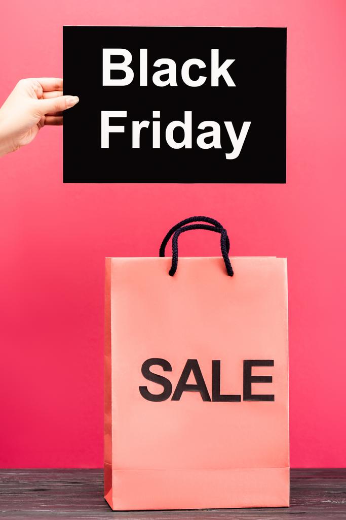 Ausgeschnittene Ansicht einer weiblichen Hand, die ein Plakat mit schwarzem Freitag-Schriftzug in der Nähe einer Einkaufstasche auf rosa hält - Foto, Bild