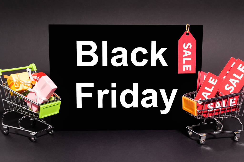 kleine Geschenke in Spielzeug-Einkaufswagen neben Plakat mit schwarzem Freitag-Schriftzug und Verkaufsanhänger auf dunklem Hintergrund  - Foto, Bild