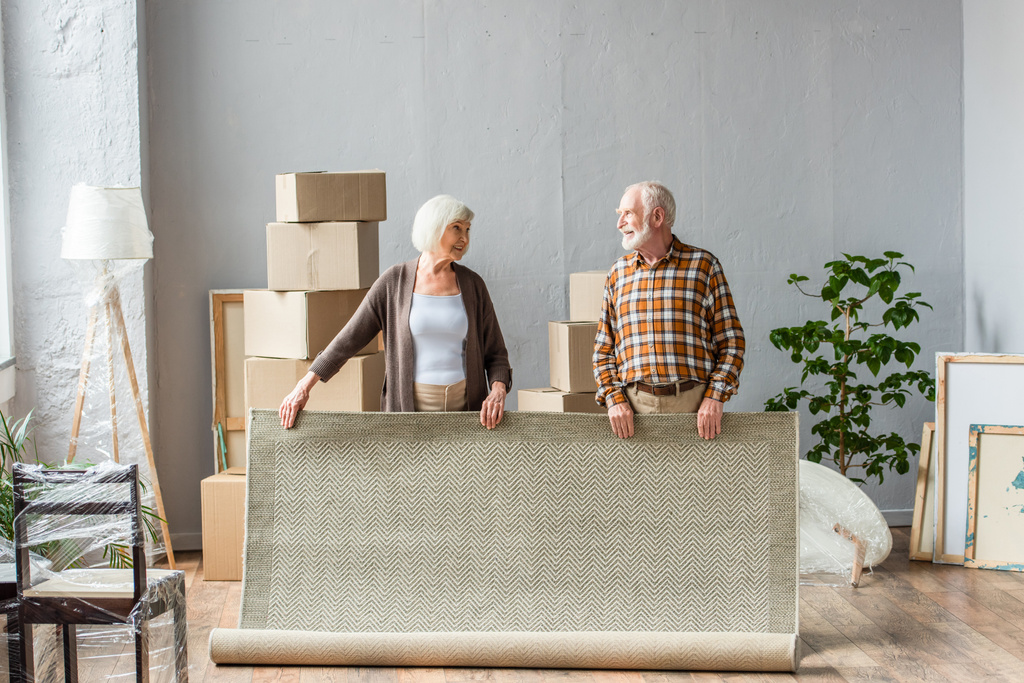 старшая пара катящийся ковер и глядя друг на друга с коробками, рамками и завод на заднем плане - Фото, изображение