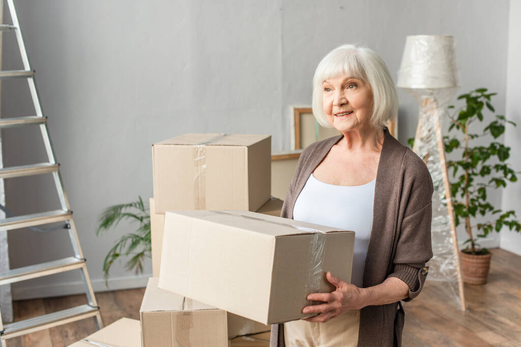 veselá starší žena držící kartónovou krabici a odvracející pohled, pohybující se koncept - Fotografie, Obrázek