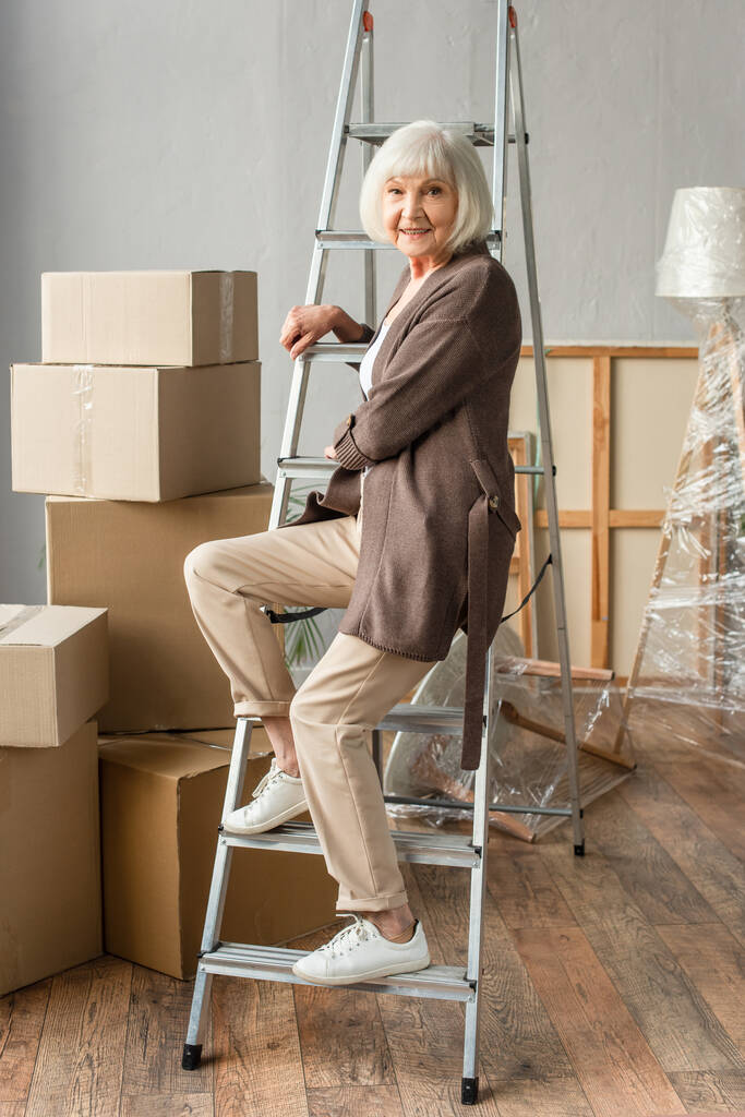 vue pleine longueur d'une femme âgée souriante assise sur une échelle avec des boîtes en carton sur le fond, concept mobile - Photo, image