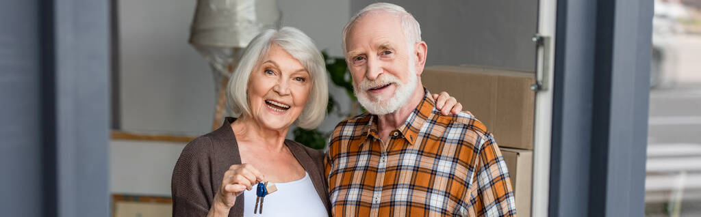 Panoramaaufnahme einer lachenden Seniorin, die Schlüssel in der Hand hält und Ehemann in neuem Haus umarmt - Foto, Bild