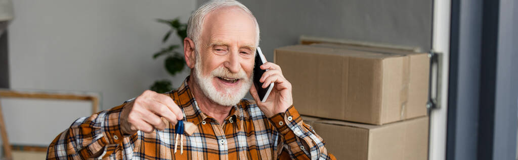 panoramiczne ujęcie szczęśliwego starszego mężczyzny rozmawiającego przez telefon i trzymającego klucze w nowym domu, poruszająca się koncepcja - Zdjęcie, obraz