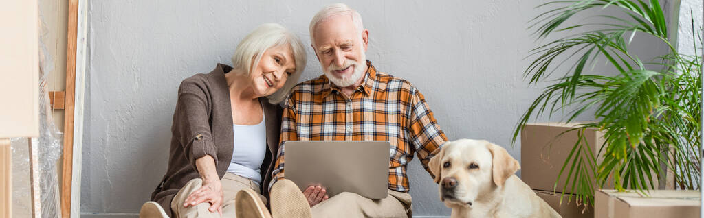 panoramisch schot van gelukkig senior paar met behulp van laptop zitten op de vloer en echtgenoot aaien hond liggen in de buurt - Foto, afbeelding