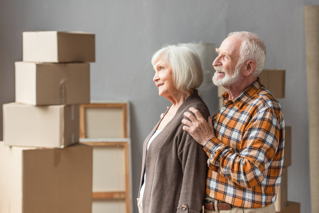cheerful husband hugging wife shoulders in new house with cardboard boxes on background - Photo, Image