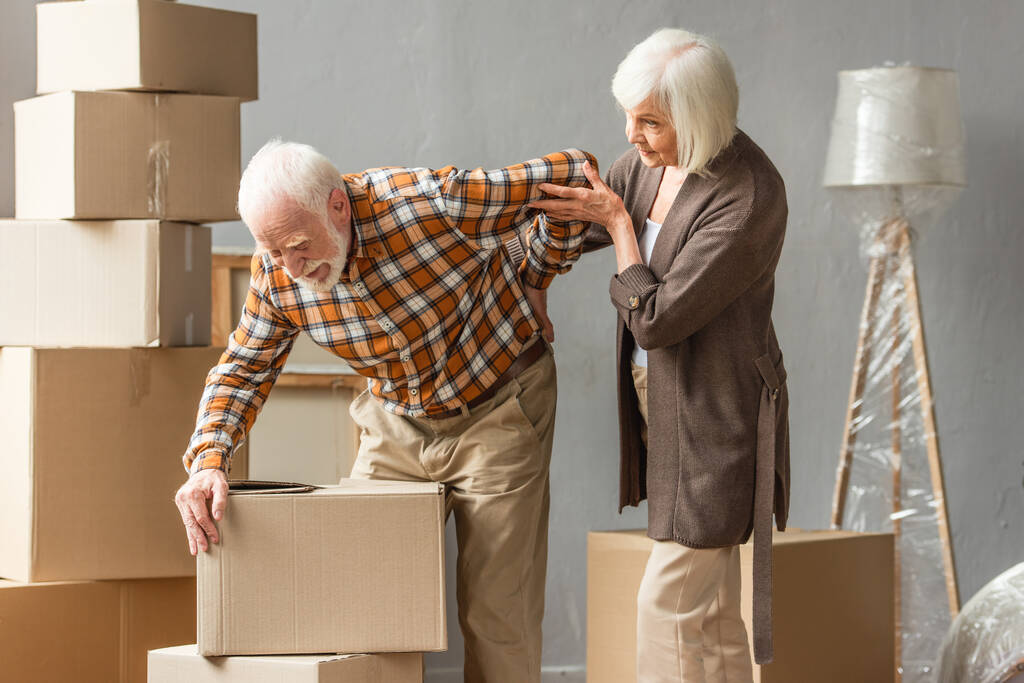 старший мужчина страдает от боли в спине и держит коробку в то время как жена пытается помочь - Фото, изображение