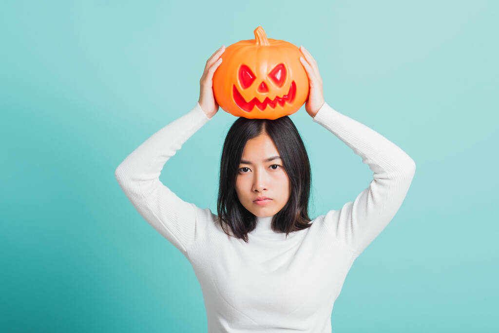 Portrait of Asian beautiful young woman holding orange model pumpkins and put it on the head, funny happy female with ghost pumpkins, studio shot isolated on blue background - Photo, Image