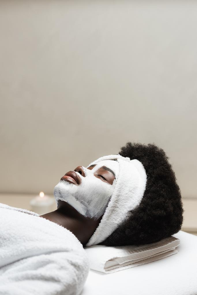 Femme afro-américaine bouclée couchée avec masque facial avec bougie floue sur fond  - Photo, image