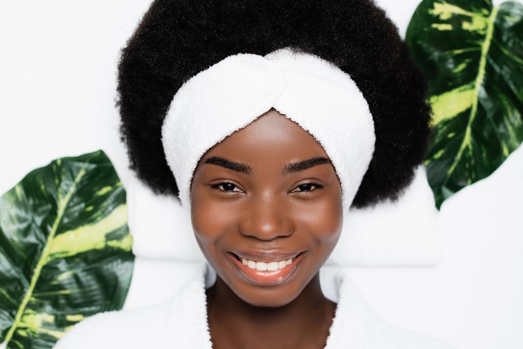 Draufsicht einer lächelnden afrikanisch-amerikanischen Frau mit weißem Stirnband, die in die Kamera blickt, während sie neben grünen Blättern im Wellness-Salon ruht - Foto, Bild