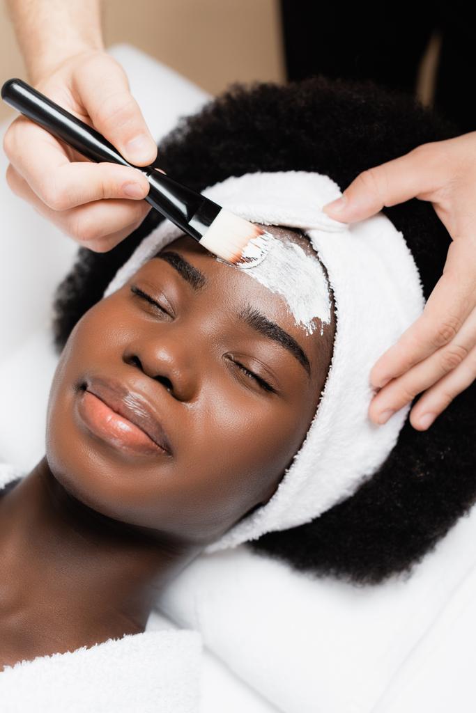 thérapeute spa appliquant masque facial avec pinceau cosmétique sur le front de la femme afro-américaine dans le salon de spa - Photo, image