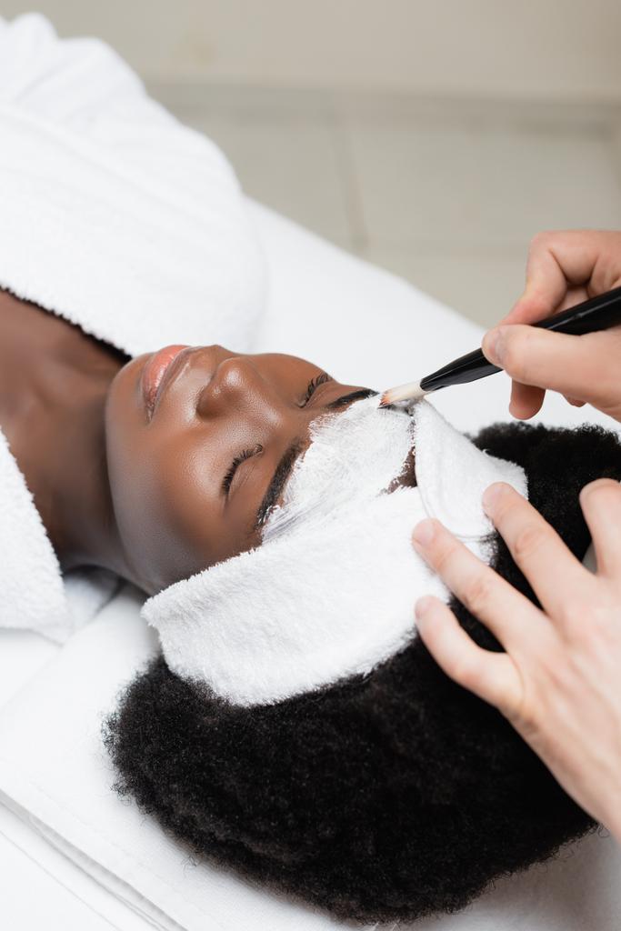 Kurtherapeutin trägt Gesichtsmaske mit kosmetischem Pinsel auf der Stirn einer Afroamerikanerin im Wellness-Salon auf - Foto, Bild