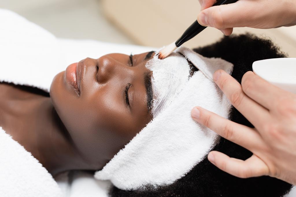 Close up view of man applying face mask with cosmetic brush on the forehead of African American woman in spa salon - Zdjęcie, obraz