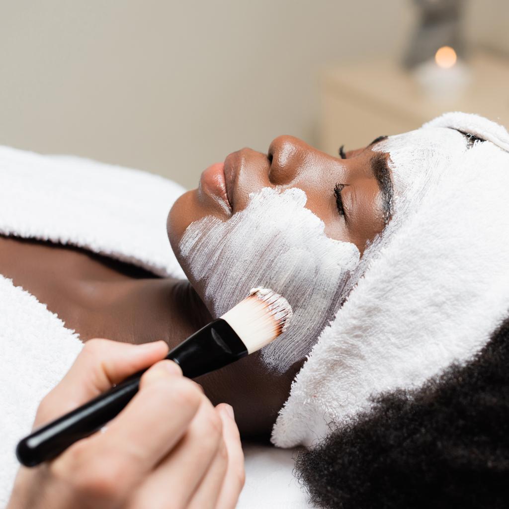 Detailní pohled lázeňského terapeuta nanášení obličejové masky s kosmetickým štětcem na tvář afroamerické ženy v lázeňském salonu na rozmazaném pozadí - Fotografie, Obrázek