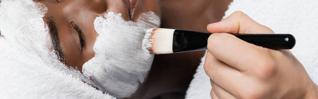 Close up άποψη του θεραπευτή spa εφαρμογή μάσκα προσώπου με καλλυντικά βούρτσα στο μάγουλο της αφρικανικής Αμερικής γυναίκα στο σαλόνι σπα, πανό - Φωτογραφία, εικόνα