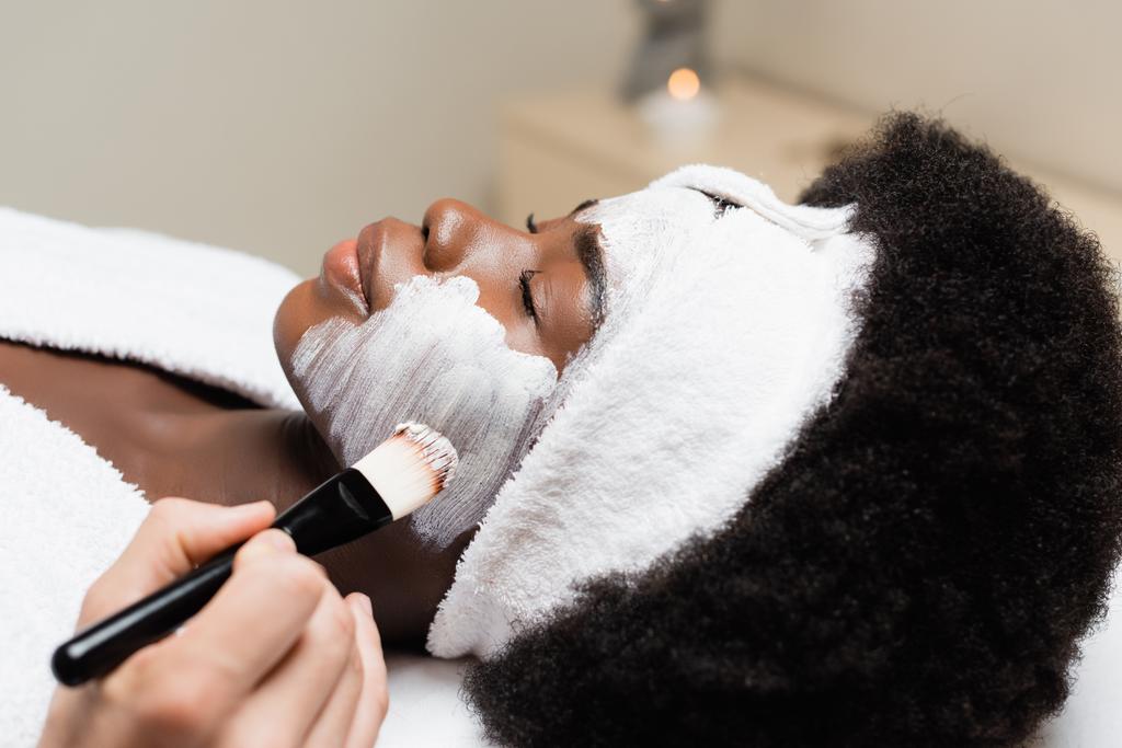 Close up widok african amerykańskiej kobiety w opasce leżącej w pobliżu mężczyzny stosowania maski twarzy na policzku w salonie spa na rozmyte tło - Zdjęcie, obraz