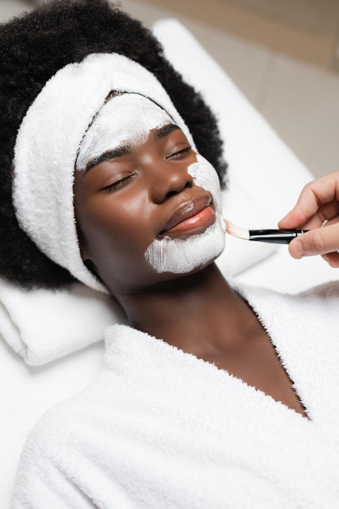 Positive afrikanisch-amerikanische Frau mit geschlossenen Augen, die in der Nähe eines Therapeuten liegt und im Wellness-Salon Gesichtsmaske am Kinn aufträgt - Foto, Bild