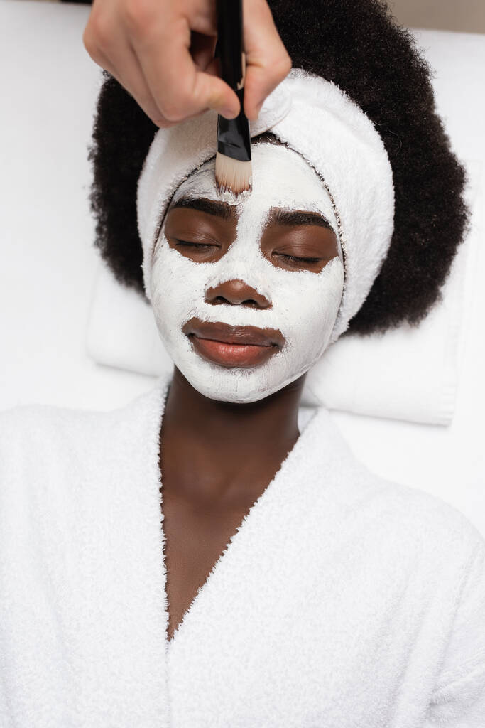 vista dall'alto della donna afro-americana positiva che indossa l'accappatoio, sdraiata vicino al terapeuta spa che applica la maschera facciale sulla fronte nel salone spa - Foto, immagini