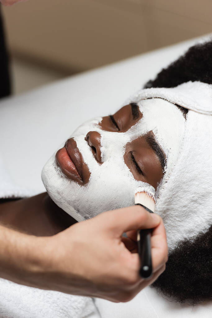 Zblízka pohled na africkou americkou ženu s čelenkou ležící poblíž lázeňské terapeutky aplikující obličejovou masku na tvář v lázeňském salonu - Fotografie, Obrázek