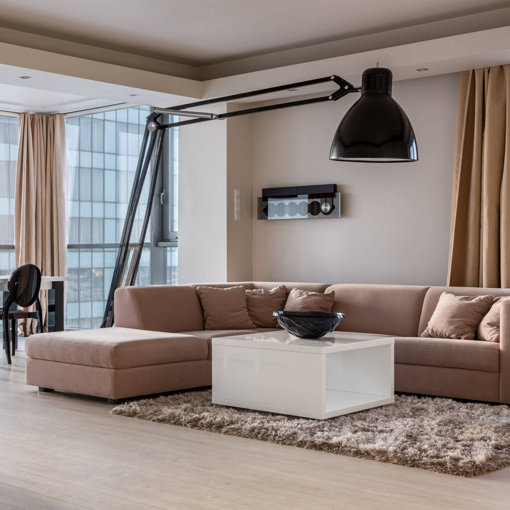 Розкішний бежевий кутовий диван у стильній вітальні з квадратним, білим журнальним столиком і сучасною великою чорною лампою
 - Фото, зображення