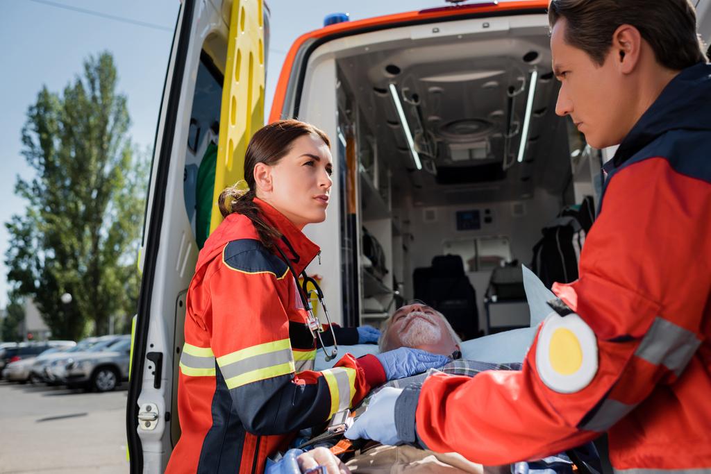 Enfoque selectivo de paramédicos en uniforme parados cerca de paciente mayor en camilla y auto ambulancia en calle urbana  - Foto, imagen