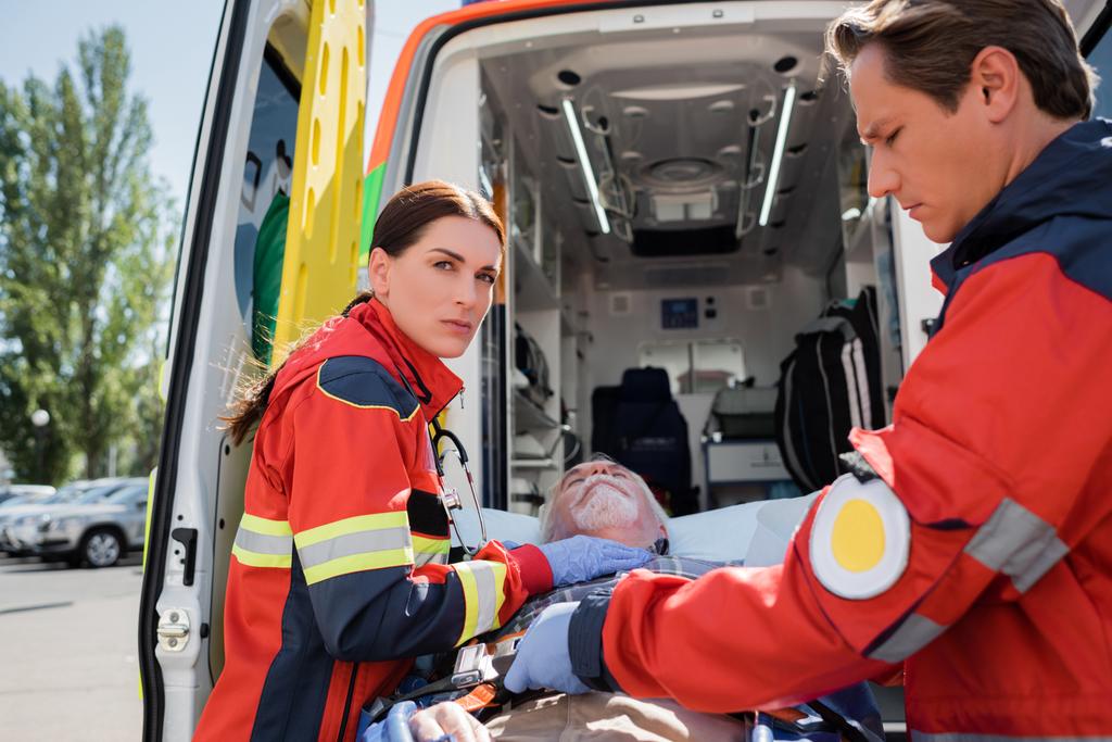 Selectieve focus van paramedicus op camera nabij patiënt op brancard en collega buiten  - Foto, afbeelding