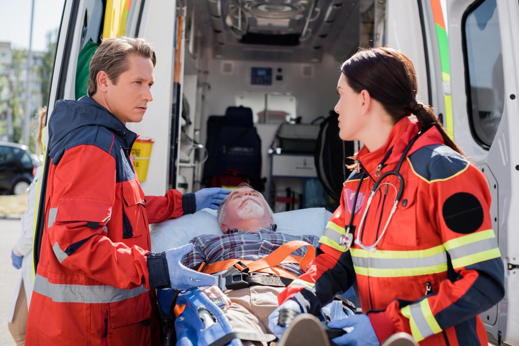Enfoque selectivo de paramédico apuntando al paciente en camilla cerca de colega en guantes de látex y coche ambulancia  - Foto, imagen