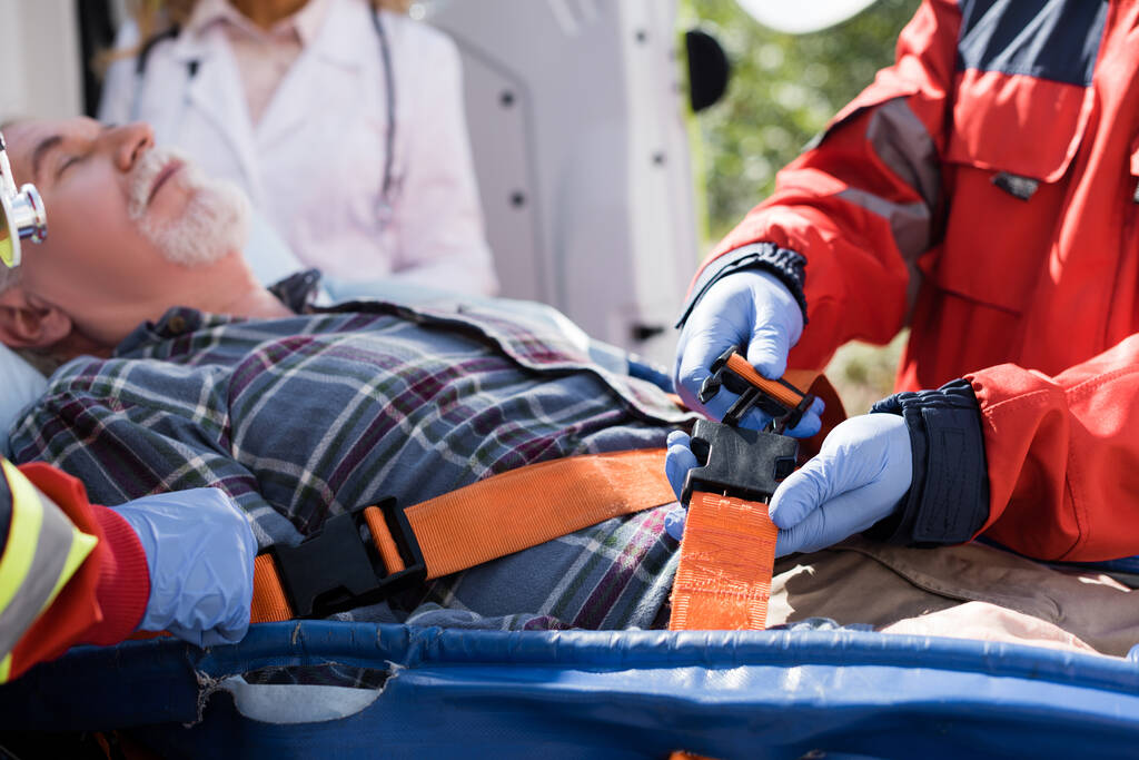 Selective focus of paramedic locking belts of stretcher near elderly patient and colleagues outdoors  - Photo, Image