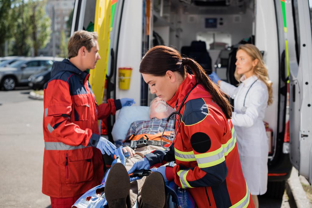 Селективный фокус пристегивания ремней скорой помощи рядом с пациентом на носилках и коллегами на городской улице  - Фото, изображение