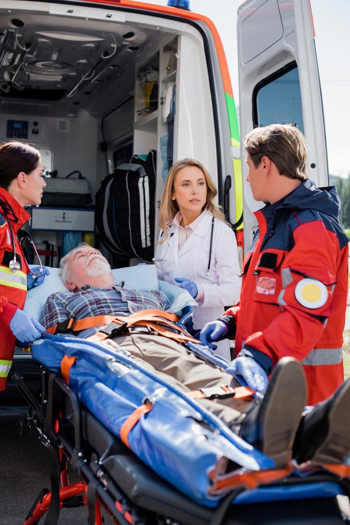 Селективный фокус врача, смотрящего на парамедика рядом с пациентом на носилках и машине скорой помощи  - Фото, изображение