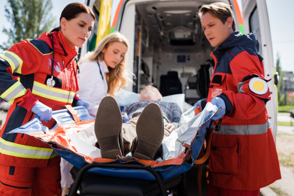Enfoque selectivo de los paramédicos que sostienen la camilla cerca del paciente, el médico y el automóvil de ambulancia al aire libre  - Foto, Imagen