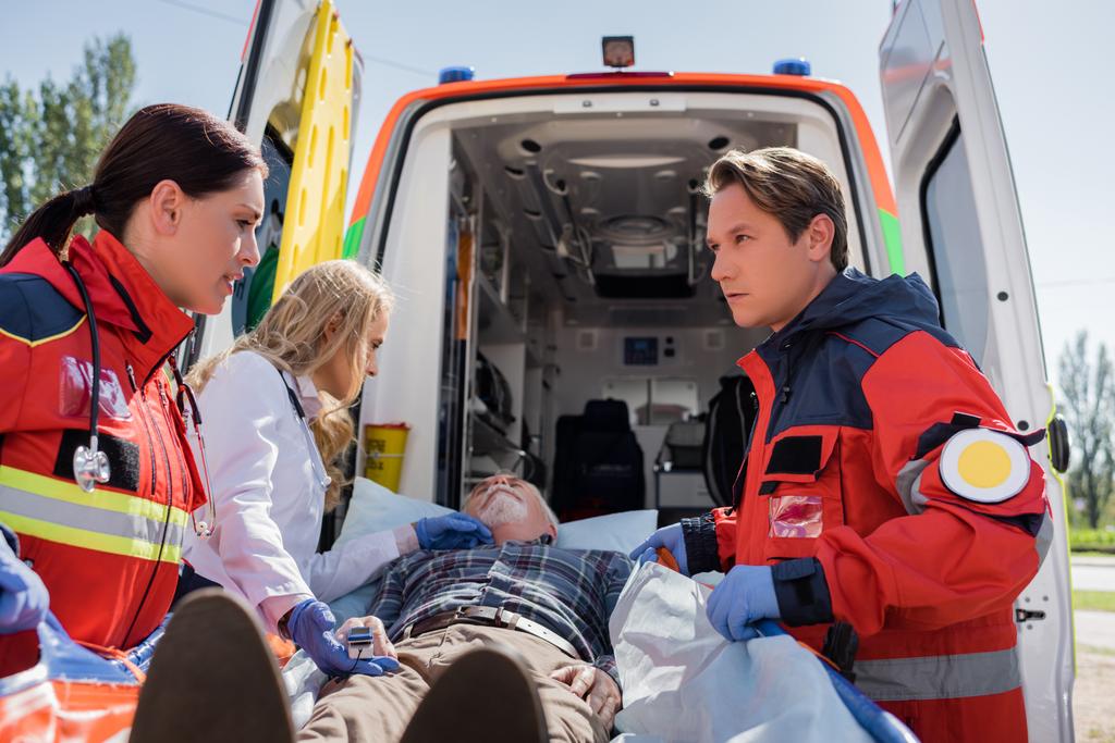 Enfoque selectivo de los paramédicos y el médico de pie cerca de hombre mayor en camilla y coche ambulancia al aire libre - Foto, imagen