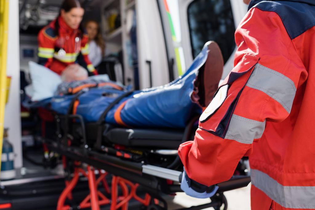Selective focus of paramedic in uniform carrying stretcher with patient in ambulance ca outdoors  - Photo, Image