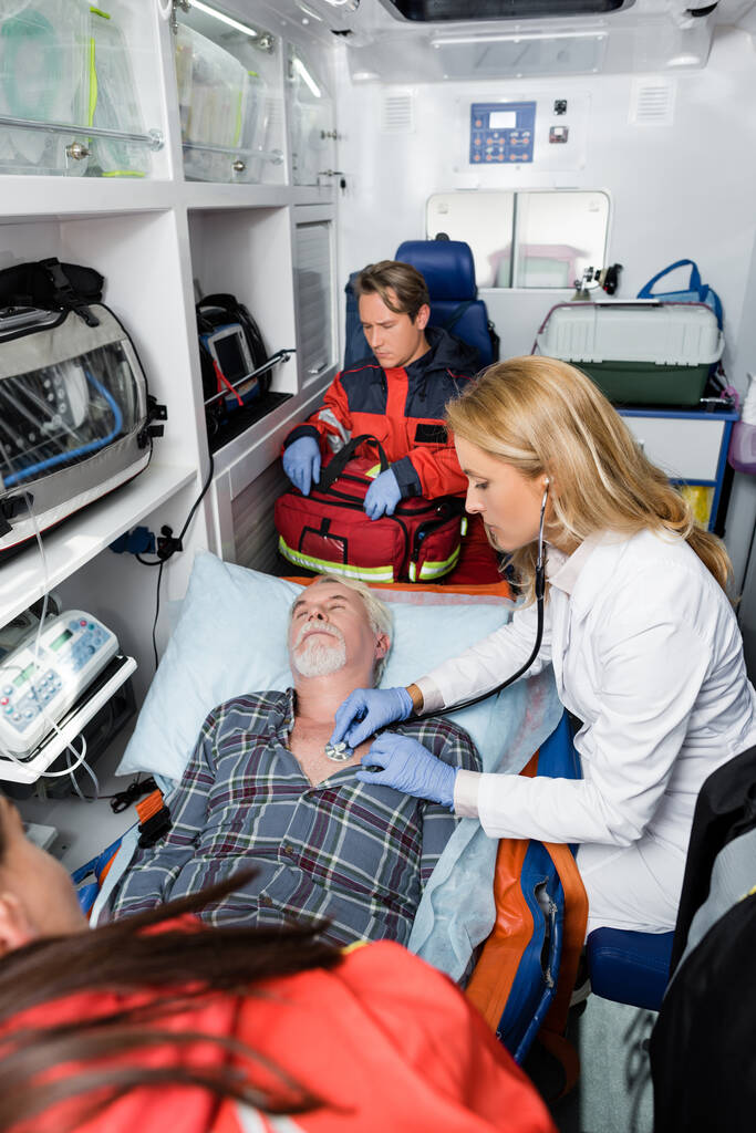 Enfoque selectivo del médico en guantes de látex examinando a paciente mayor con estetoscopio cerca de paramédicos en uniforme en coche ambulancia  - Foto, Imagen