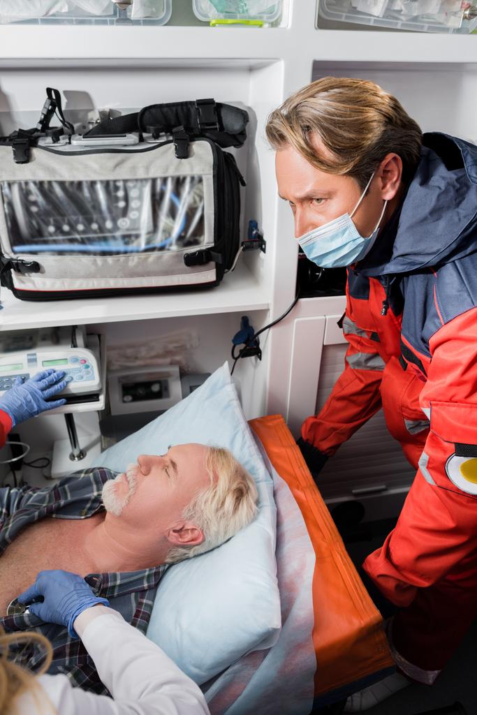 Ensihoitajan valikoiva painopiste lääketieteellisessä naamiossa, joka seisoo lähellä lääkäriä, joka tutkii iäkkäitä potilaita stetoskoopilla ambulanssiautossa   - Valokuva, kuva
