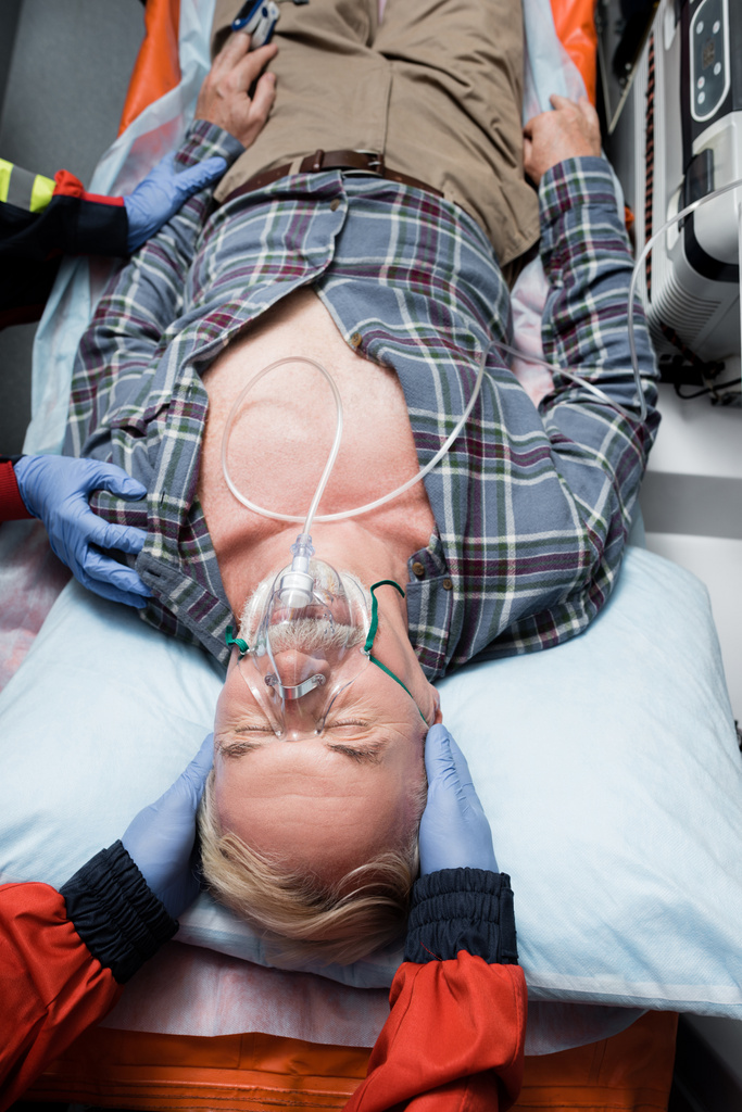 Вид пациента в кислородной маске, лежащего рядом с парамедиками в латексных перчатках в машине скорой помощи  - Фото, изображение