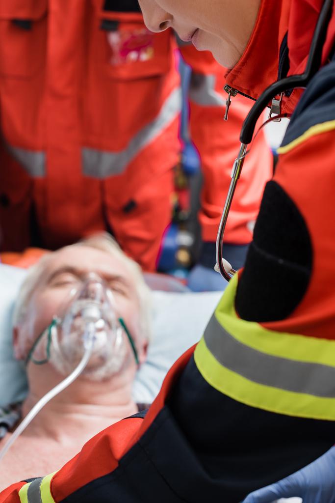 Selective focus of paramedic in uniform standing near elderly man in oxygen mask during first aid  - Photo, Image