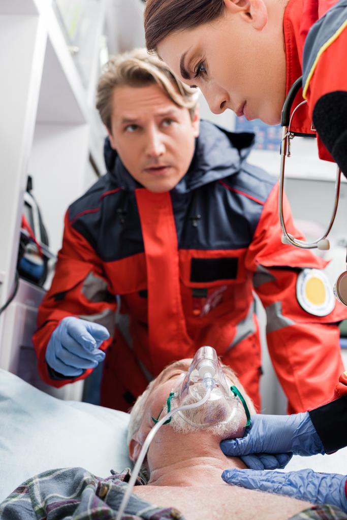 Selektivní zaměření záchranné kontroly pulsu pacienta v kyslíkové masce v blízkosti kolegy v sanitce auto  - Fotografie, Obrázek
