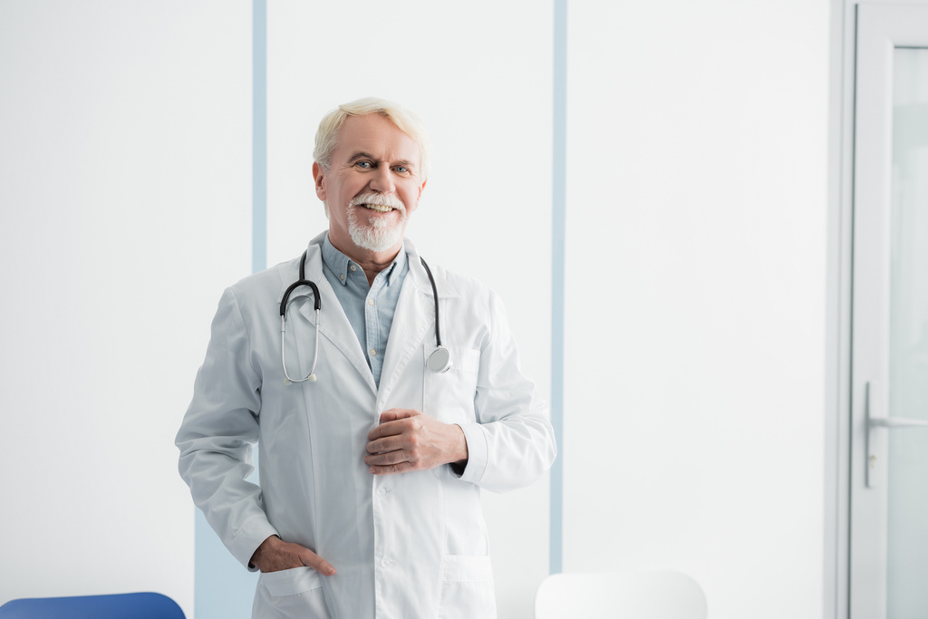 Starszy lekarz z ręką w kieszeni patrzący na kamerę w szpitalu  - Zdjęcie, obraz
