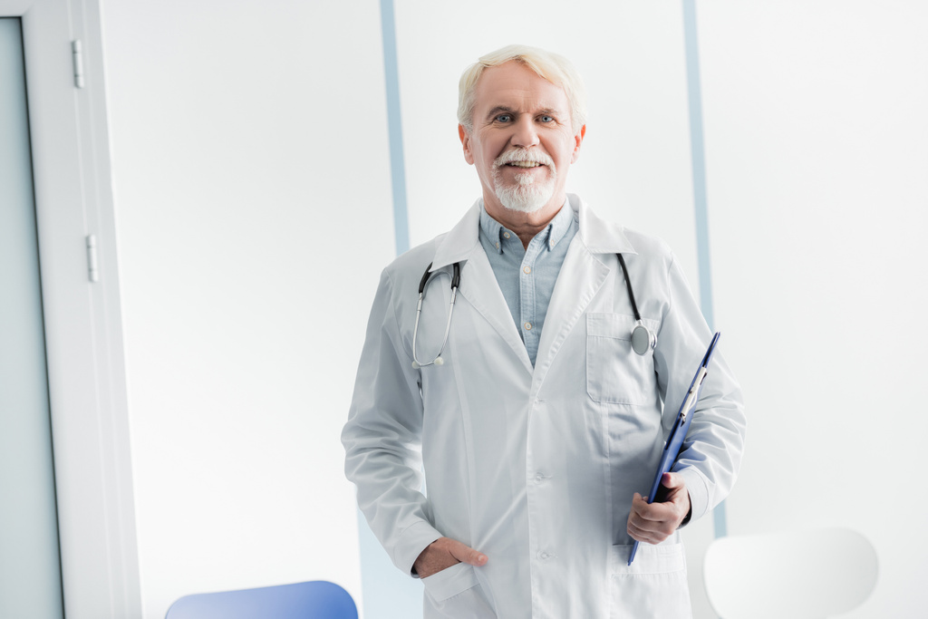 Médico de cabelos grisalhos com mão no bolso segurando prancheta no hospital  - Foto, Imagem