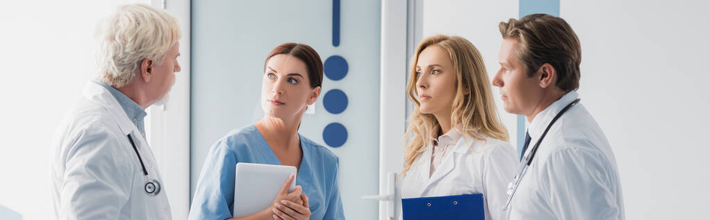 Zdjęcie panoramiczne lekarzy i pielęgniarek z tabletem cyfrowym patrzących na starszego kolegę w klinice   - Zdjęcie, obraz