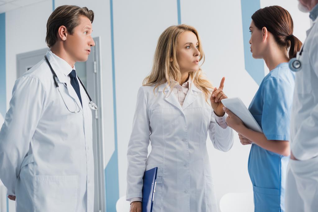 Selektywne skupienie się lekarza ze schowkiem wskazującym palcem w pobliżu kolegów i pielęgniarki z tabletem cyfrowym  - Zdjęcie, obraz