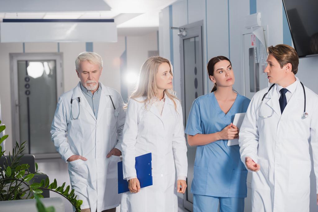 Concentration sélective du médecin parlant tout en marchant près de collègues avec presse-papiers et tablette numérique à l'hôpital  - Photo, image
