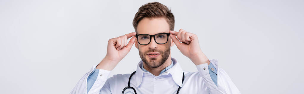 Retrato de un médico adulto joven que sostiene el marco de las gafas, mientras mira a la cámara aislada en blanco, pancarta - Foto, Imagen
