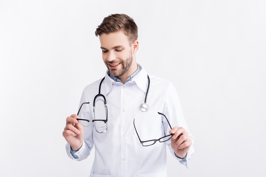 Médico sorridente com estetoscópio segurando par de óculos com molduras pretas isoladas em branco - Foto, Imagem