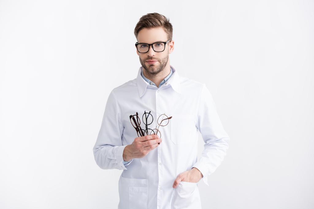 Vue de face du jeune ophtalmologiste adulte avec la main dans la poche tenant plusieurs lunettes isolées en blanc - Photo, image