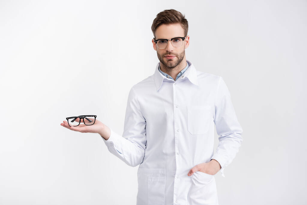 Vista frontale del medico fiducioso con mano in tasca che tiene gli occhiali sul palmo della mano, mentre guarda la fotocamera isolata su bianco - Foto, immagini