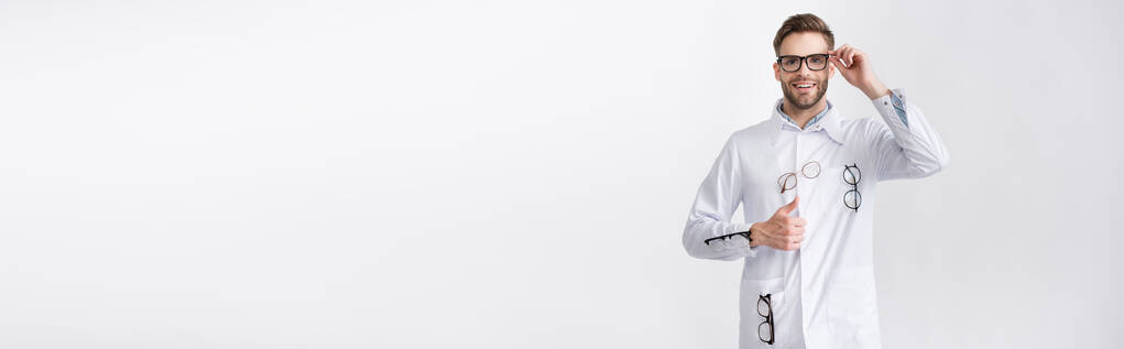 Vooraanzicht van blije oogarts met duim omhoog, in witte jas met hangende brillen geïsoleerd op wit, spandoek - Foto, afbeelding