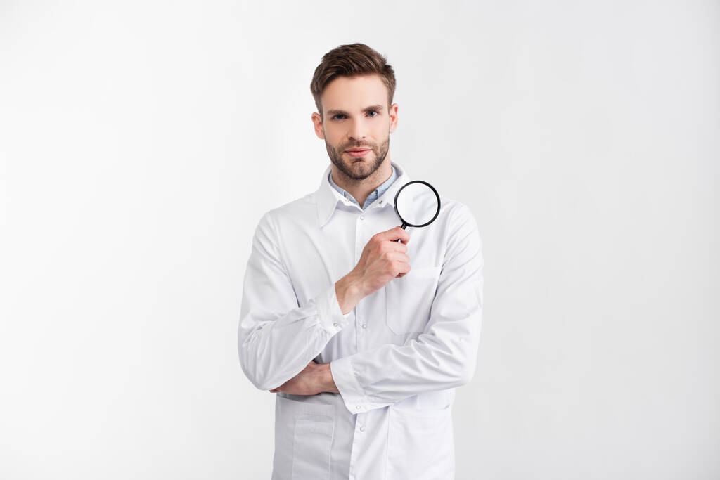 Přední pohled na oftalmologa v bílém plášti s lupem při pohledu na kameru izolovanou na bílém - Fotografie, Obrázek