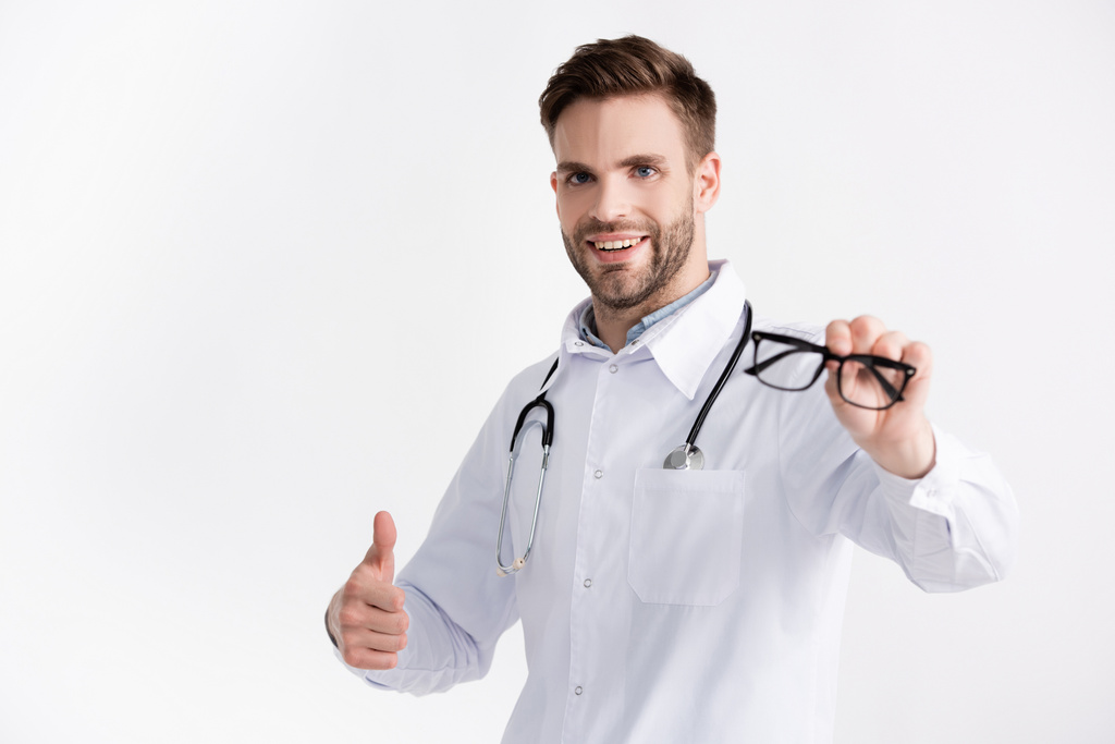 Glimlachende oogarts met stethoscoop, met goedkeuring gebaar, terwijl het tonen van bril op wazig voorgrond geïsoleerd op wit - Foto, afbeelding