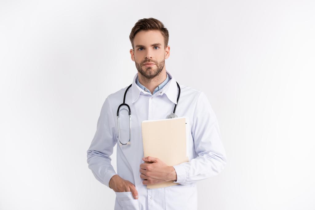 Frontansicht des selbstbewussten Arztes mit einer Hand in der Tasche, die einen Ordner hält, während er die Kamera isoliert auf Weiß betrachtet - Foto, Bild